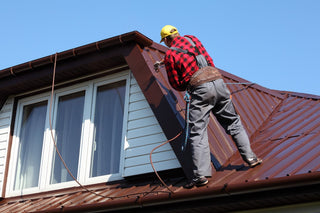 Homeowner in safety gear using a hose to clean a metal roof, showcasing effective maintenance on a sunny day with a garden in the background.